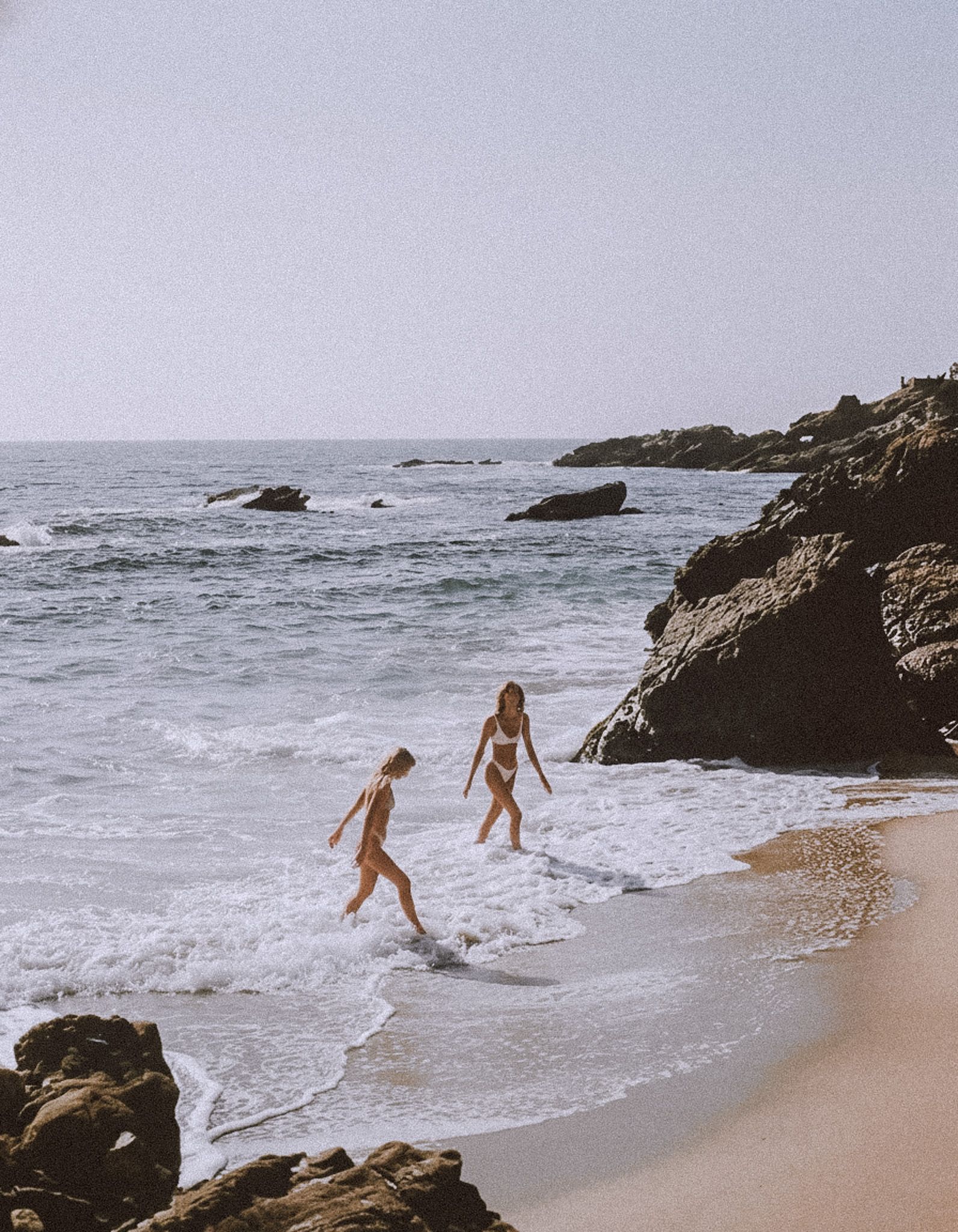 Two women walking into the shore break below a headland